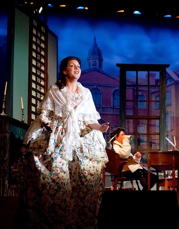 1776 — a woman in an elaborate floral colonial gown sings a heartfelt solo before the voting board and a Philadelphia backdrop while a man writes at a desk behind her