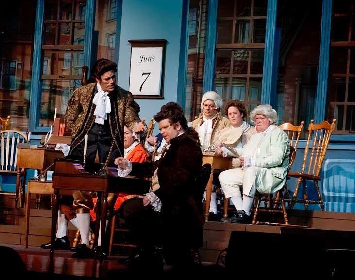 1776 — three actors in colonial period costumes perform a dramatic scene on stage before the Philadelphia backdrop