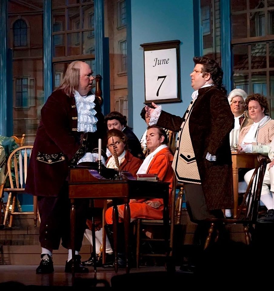 1776 — Benjamin Franklin in a burgundy coat faces John Adams in a dark brown coat across a writing desk as the full Congress looks on, a June 7 calendar on the wall behind them