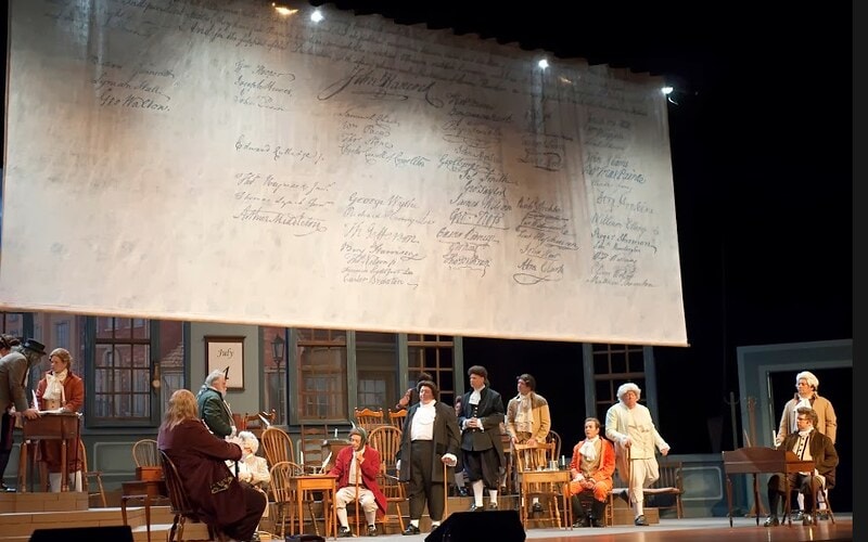 1776 — the full ensemble of delegates stands before a giant projected Declaration of Independence backdrop bearing all the signatories' signatures, a July calendar visible at lower left