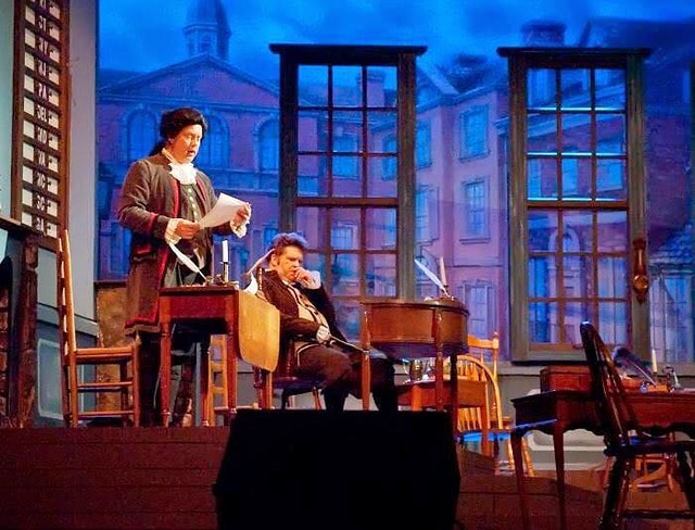 1776 — two performers in colonial costume stand together before the Philadelphia backdrop in a quiet two-person scene under warm stage lighting