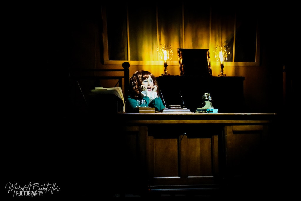 Annie — young Annie sits alone at a large wooden desk in a dramatic golden spotlight