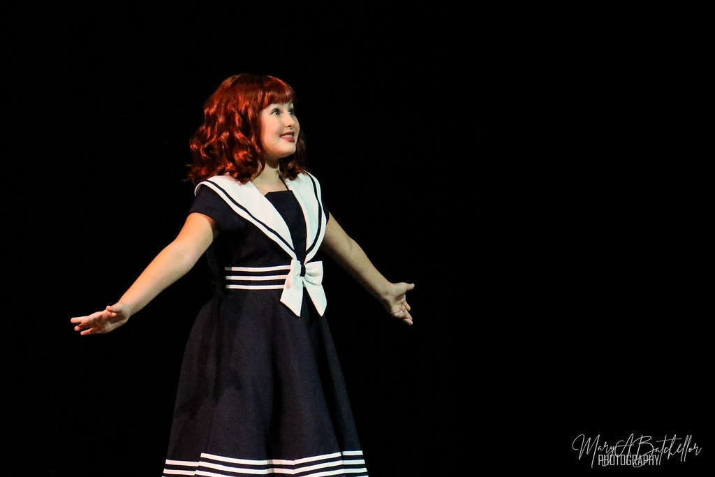 Annie — Annie in a navy sailor dress sings a solo in a dramatic spotlight
