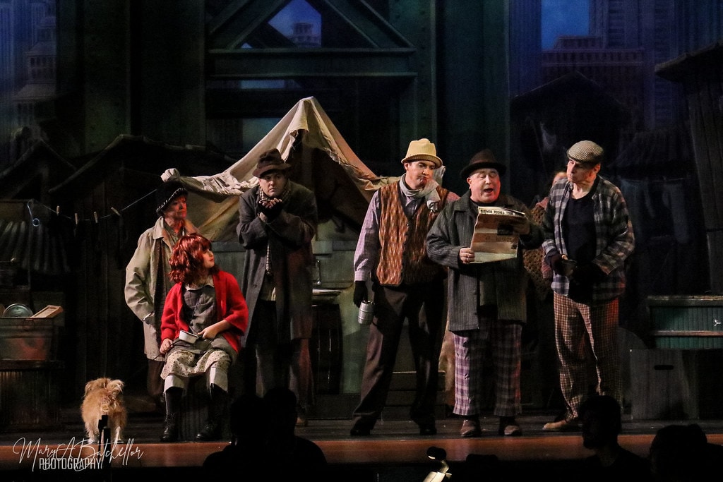 Annie — Annie sits among five men in Depression-era coats beside a tent in the Hooverville set