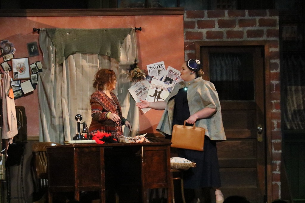 Annie — Miss Hannigan faces a woman in a navy cape at the orphanage office desk — wider angle