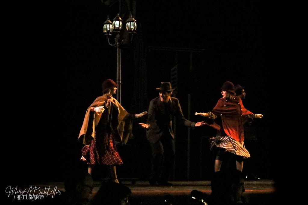 Annie — three performers in Depression-era capes and fedoras dance around a vintage street lamp