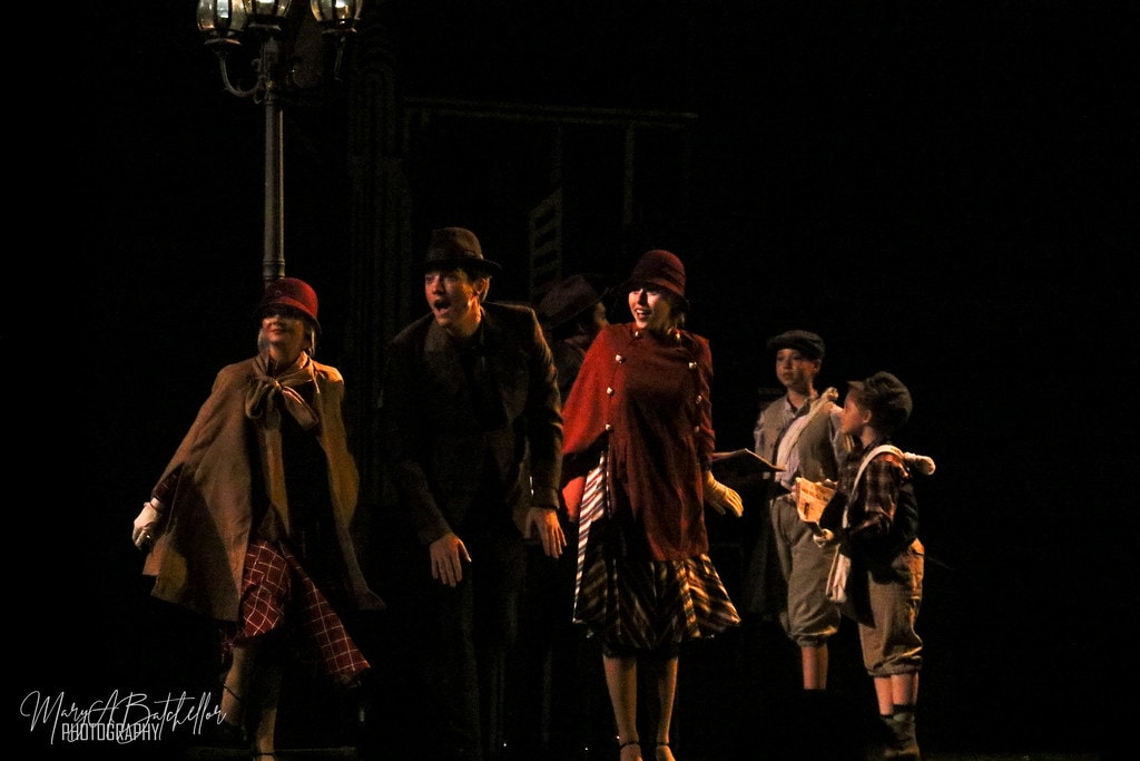 Annie — five performers in Depression-era coats walk and sing together under a vintage street lamp