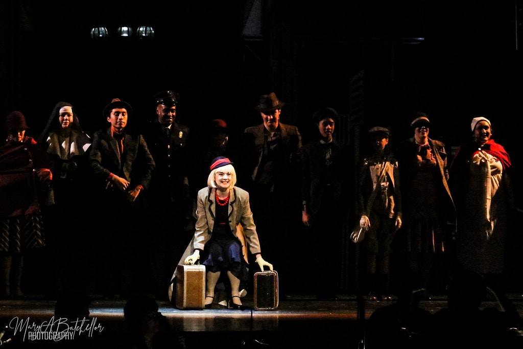 Annie — Grace Farrell kneels center stage with two suitcases as the full ensemble stands behind her
