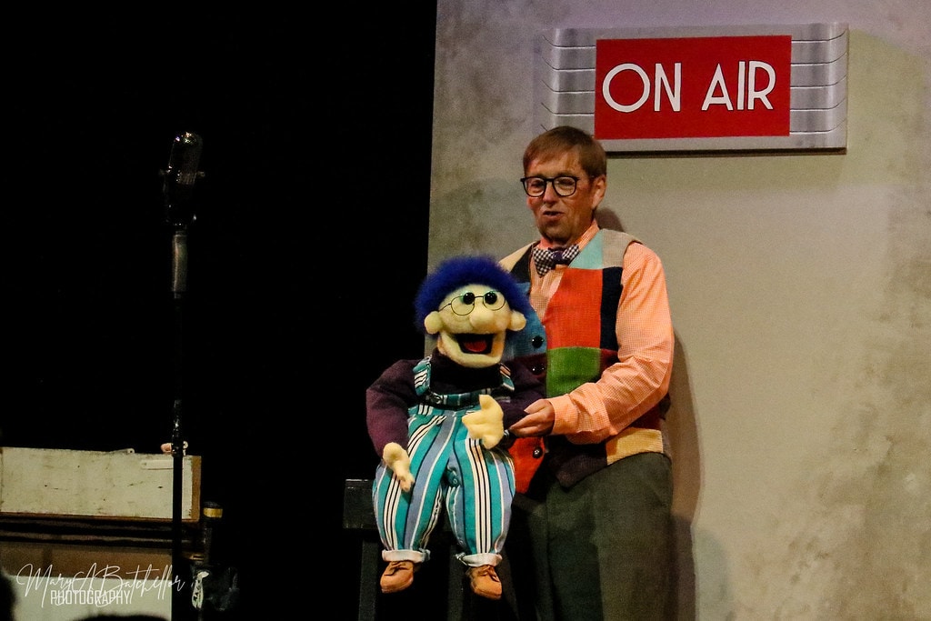 Annie — the ventriloquist in a colorful patchwork vest holds his blue-haired puppet beside a vintage microphone