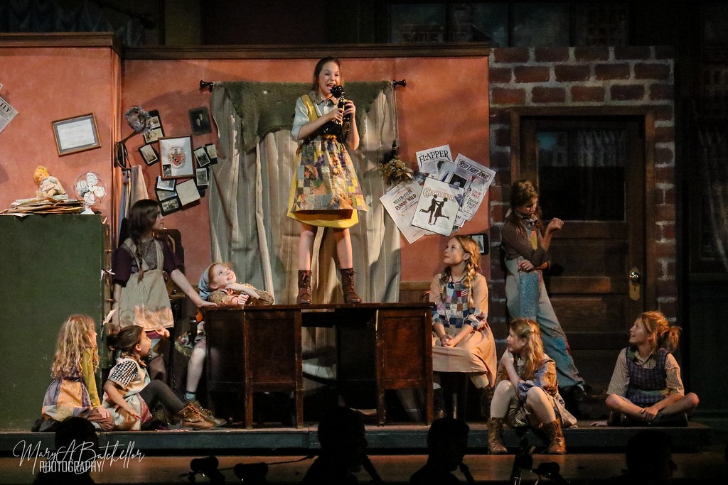 Annie — Annie on a wooden cabinet holding a vintage telephone as nine orphan girls watch