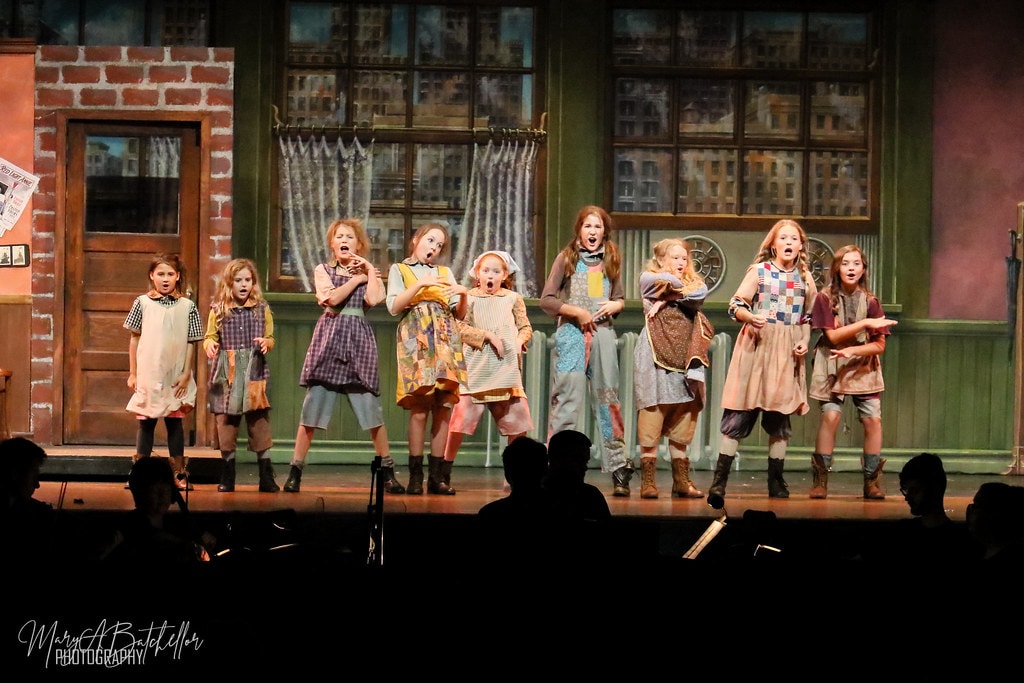 Annie — nine orphan girls react with wide-open mouths in a powerful ensemble moment