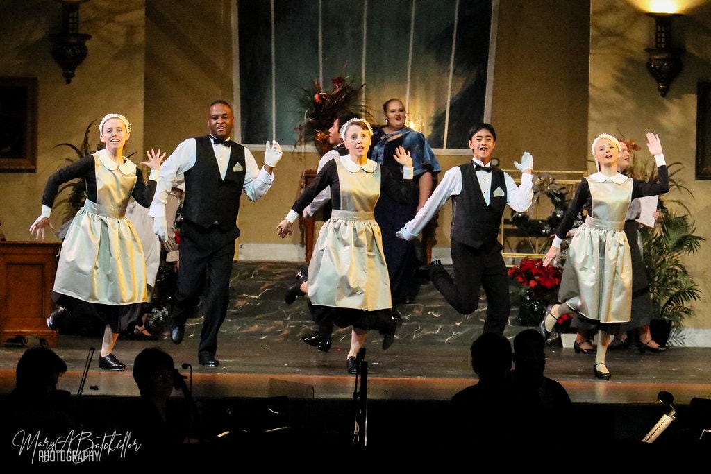 Annie — six Warbucks mansion servants dance with raised hands before the Christmas-decorated ballroom