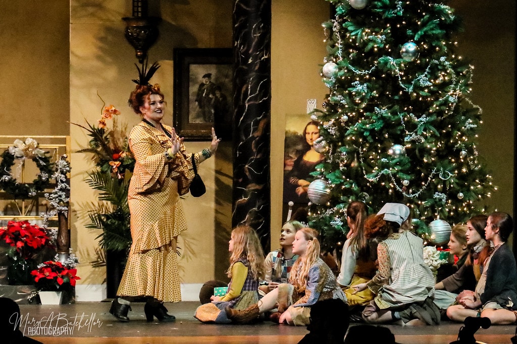 Annie — Miss Hannigan gestures dramatically as seven orphan girls sit at her feet beside the Christmas tree