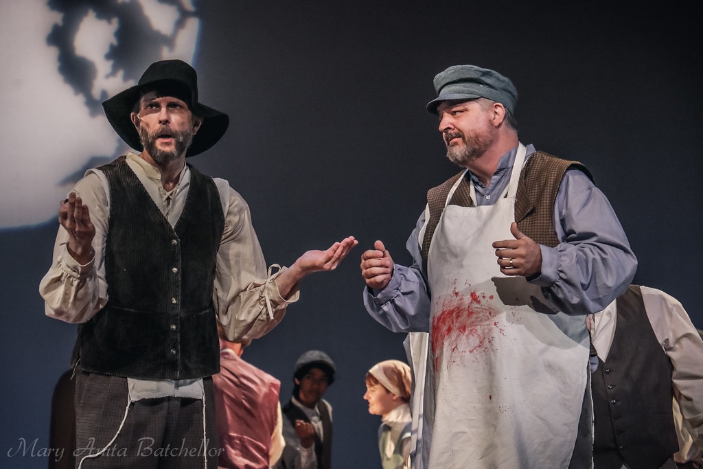 Fiddler on the Roof — the Rabbi in a black hat and the Butcher in a bloodied apron face each other in a dramatic scene before the moon backdrop