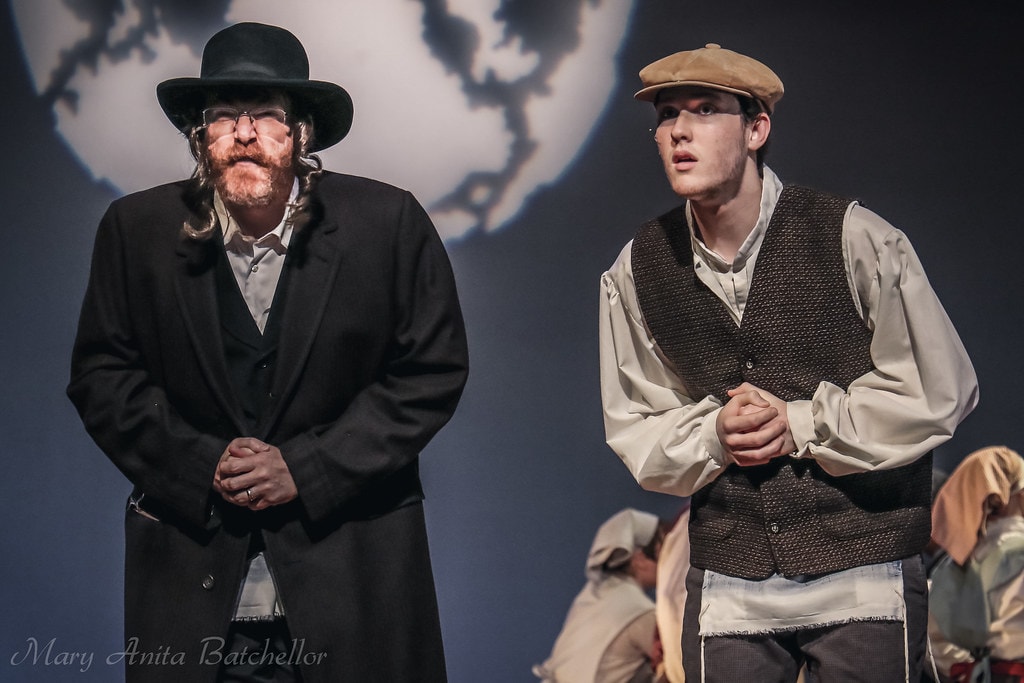 Fiddler on the Roof — the Rabbi in a long black coat and a young man in a newsboy cap and vest stand solemnly before the large moon backdrop