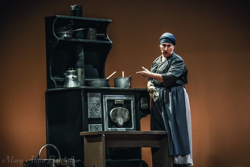 Fiddler on the Roof — Yente the Matchmaker in a blue headscarf gestures dramatically at her wood-burning stove in the kitchen scene