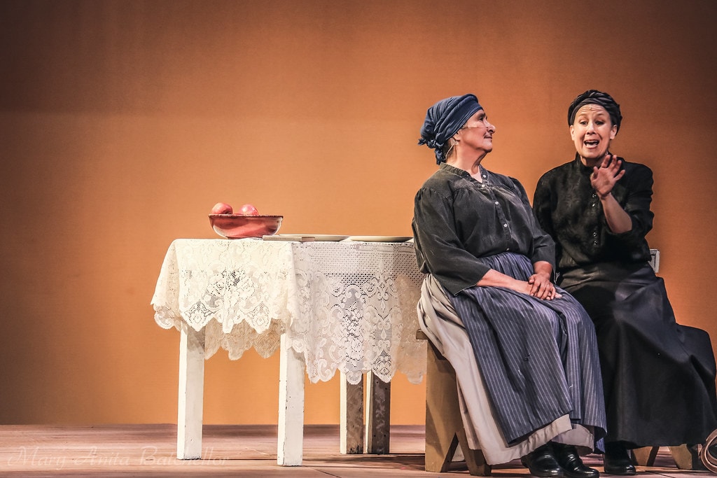 Fiddler on the Roof — Golde and Yente sit together at a lace-covered table with a bowl of apples, gossiping in the warm kitchen scene