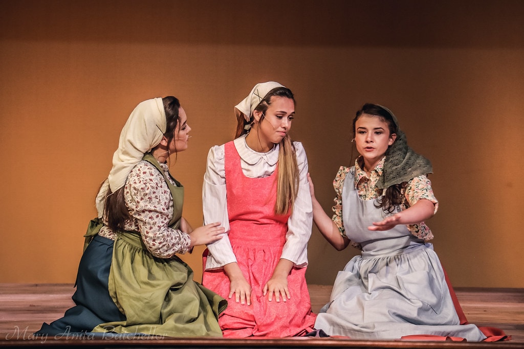 Fiddler on the Roof — three of Tevye's daughters in aprons and headscarves kneel together in an emotional scene on the warm amber-lit stage