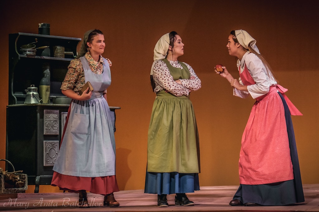 Fiddler on the Roof — three daughters in aprons and headscarves converse by the kitchen stove, one holding an apple in the warm amber kitchen set
