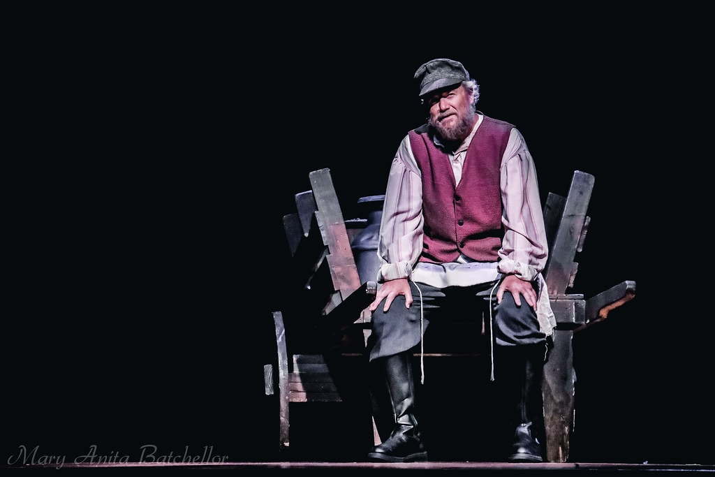 Fiddler on the Roof — Tevye in a red vest and newsboy cap sits alone on a wooden cart bench in a dramatic spotlight against a black backdrop