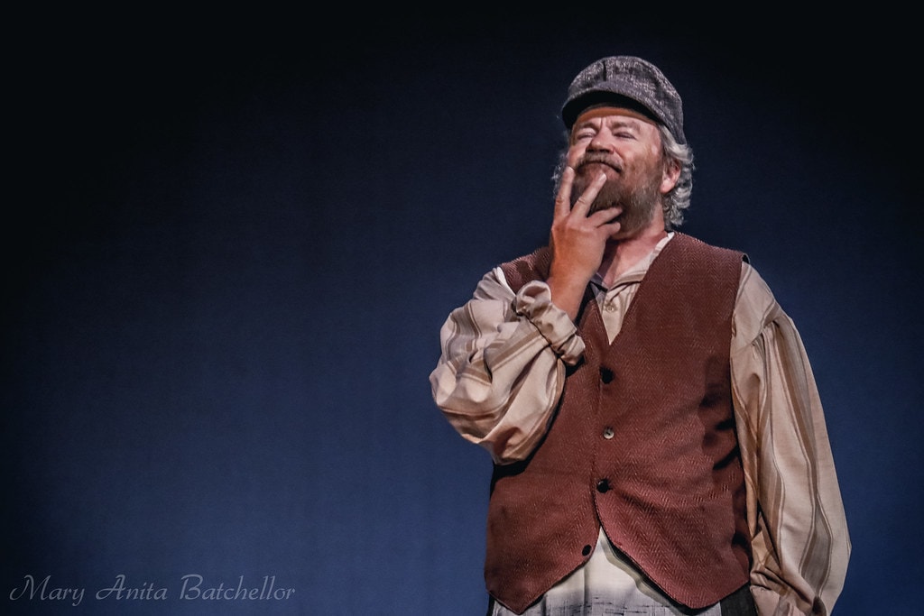 Fiddler on the Roof — Tevye in a red-brown vest and newsboy cap strokes his beard thoughtfully in a close dramatic portrait against a blue backdrop