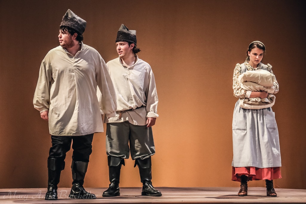 Fiddler on the Roof — two young men in fur caps and white shirts stand beside a young woman in a floral blouse and apron holding bundled linens against the warm amber backdrop