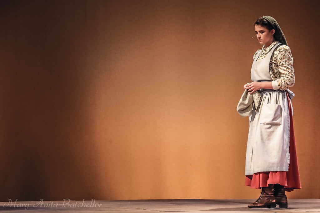 Fiddler on the Roof — a young woman in a floral blouse, white apron, and red skirt stands alone in a solitary spotlight against the warm amber backdrop