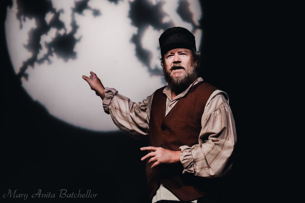 Fiddler on the Roof — Tevye in a brown vest and plaid shirt sings a dramatic solo before the large moon backdrop