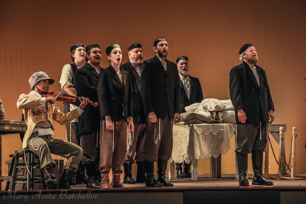 Fiddler on the Roof — the Fiddler plays violin seated at left while the full male ensemble in dark coats and caps sings together beside a lace-covered Sabbath table