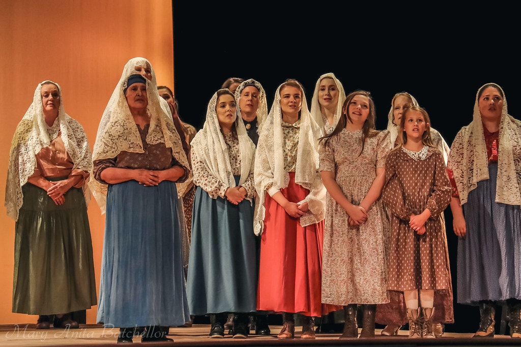 Fiddler on the Roof — the full female ensemble of women and girls in headscarves and long skirts sings together against the warm amber and black backdrop