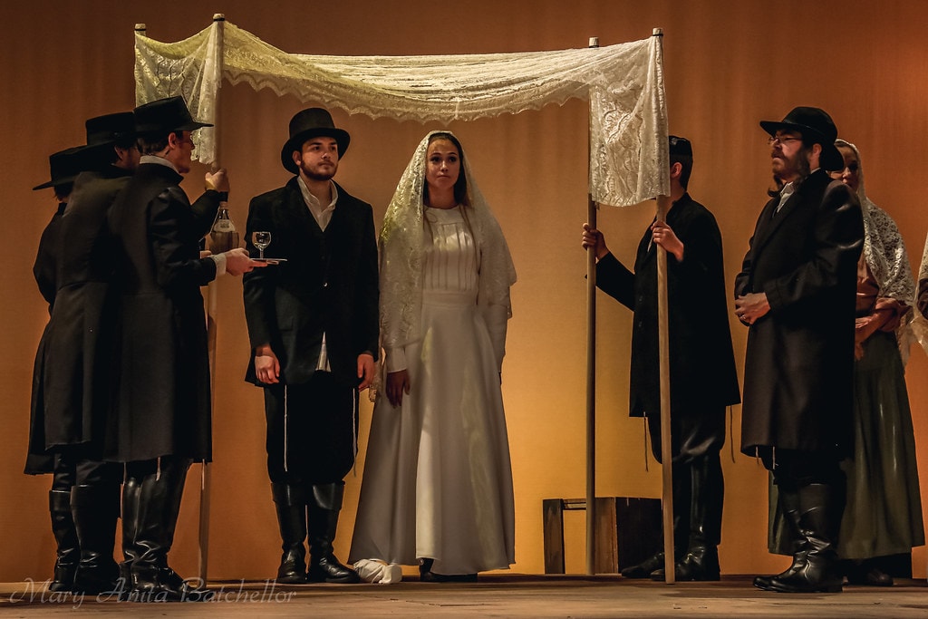 Fiddler on the Roof — a bride in a white gown and veil stands solemnly under a lace wedding canopy flanked by men in black coats and top hats in the wedding ceremony scene