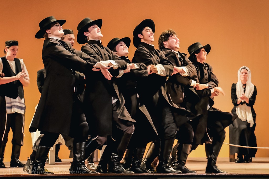 Fiddler on the Roof — a line of seven men in black coats and wide-brimmed hats lean back laughing and dancing together in the joyful wedding celebration number