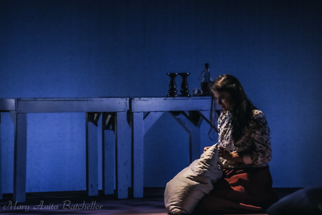 Fiddler on the Roof — a young woman in a floral blouse and red skirt sits alone on the floor beside a white table with candlesticks in a quiet blue-lit solitary scene