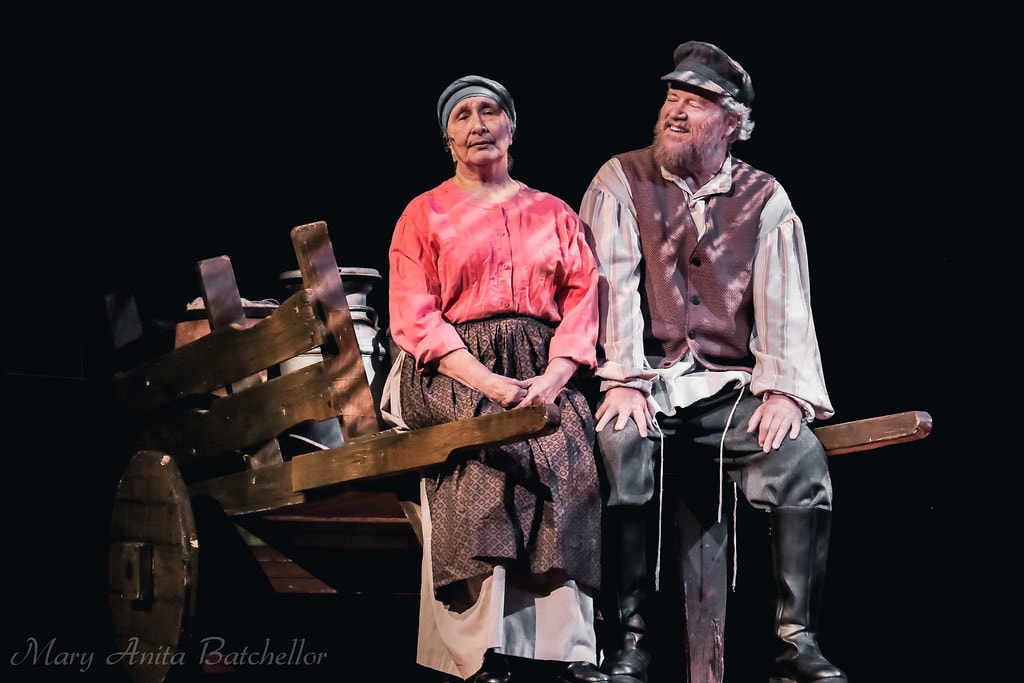 Fiddler on the Roof — Golde in a pink blouse and Tevye in a brown vest sit together on the wooden milk cart laughing warmly in a tender scene against a black backdrop
