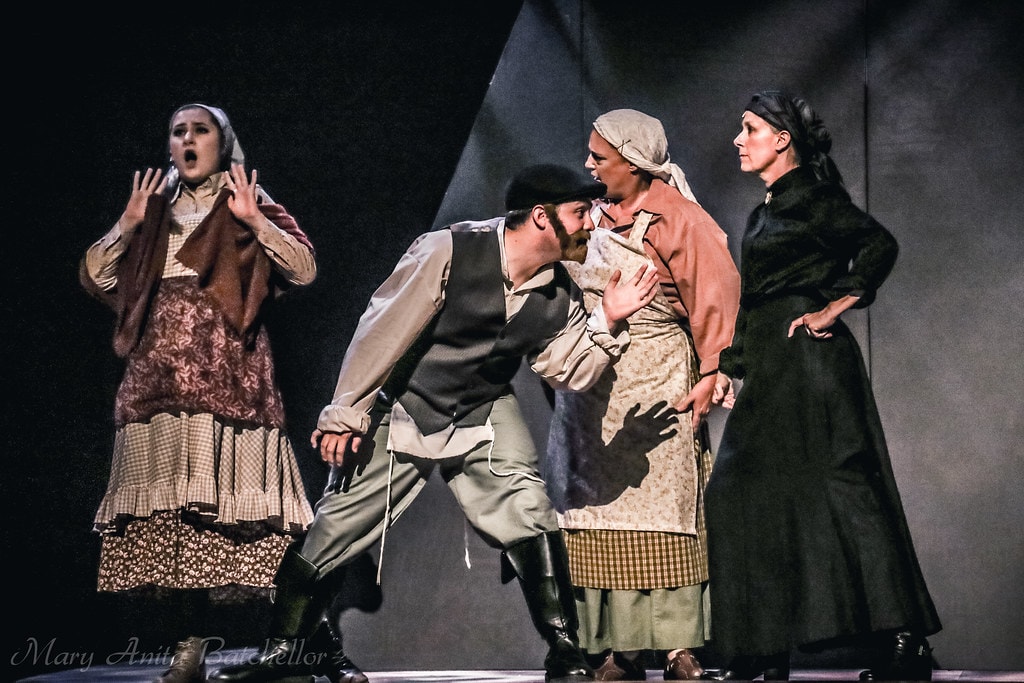 Fiddler on the Roof — a young woman in a floral dress reacts with raised hands as a man in a vest leans toward another woman while a stern woman in black looks on in a dramatic confrontation scene