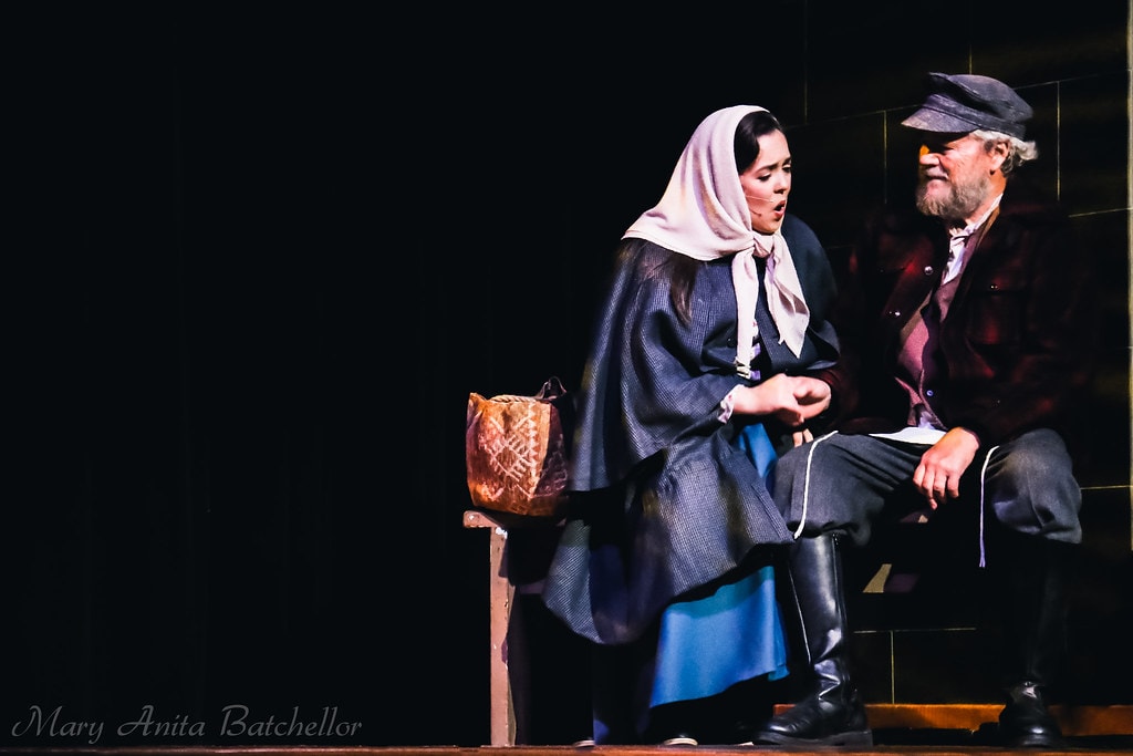 Fiddler on the Roof — a young woman in a blue headscarf and cape sits on a wooden bench beside Tevye in a red plaid shirt in a quiet intimate scene against a black backdrop