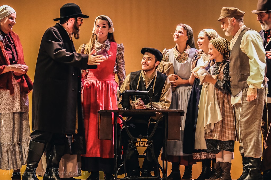 Fiddler on the Roof — the full ensemble of villagers in period costumes gathers warmly around a young man seated at a sewing machine in a joyful amber-lit scene