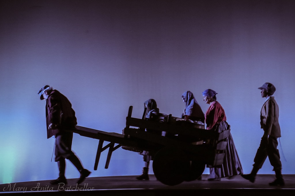 Fiddler on the Roof — five villagers in period costumes push and walk alongside a wooden cart in a haunting blue-lit silhouette scene representing the expulsion from Anatevka