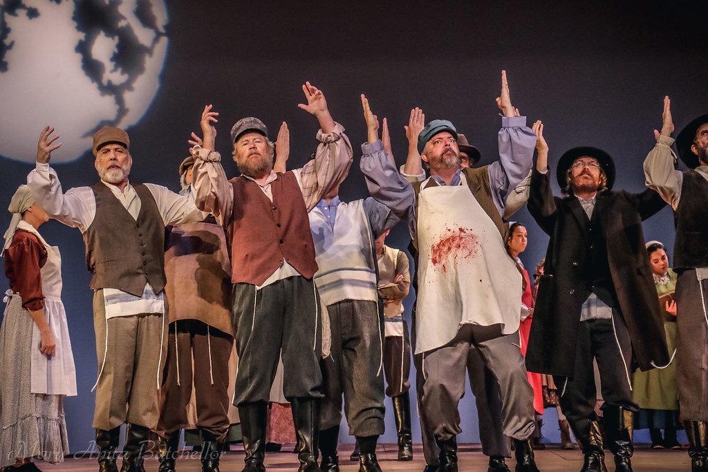 Fiddler on the Roof — the full male ensemble raises their hands skyward in a powerful ensemble moment before the moon backdrop