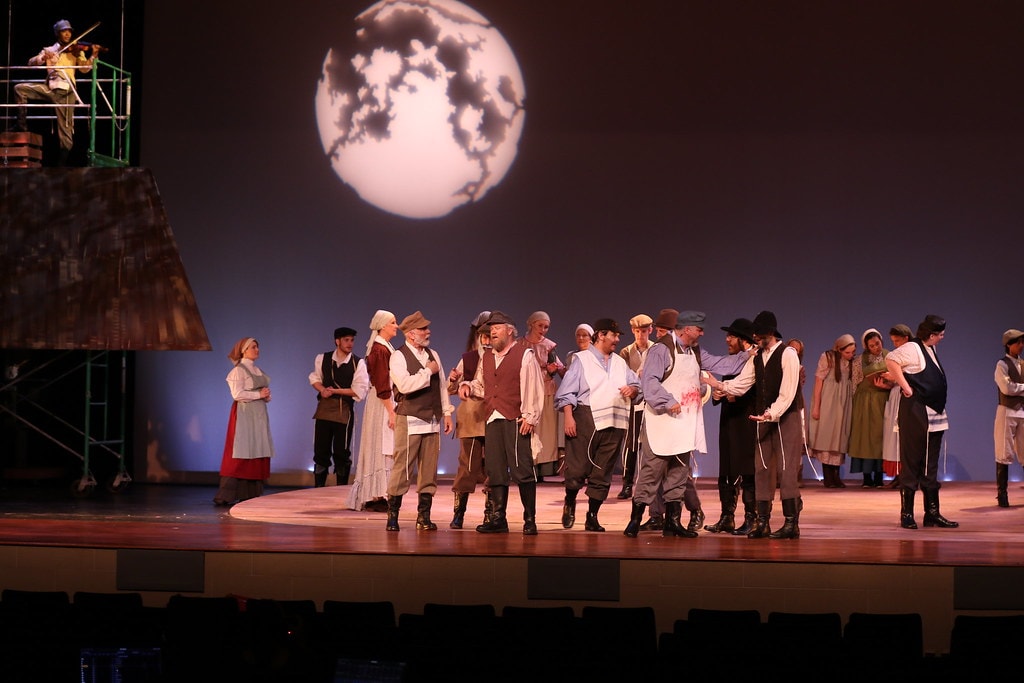 Fiddler on the Roof — the full mixed ensemble of villagers in period costumes gathers on the warm wooden stage floor before the large glowing moon backdrop, the Fiddler visible on the rooftop scaffolding at upper left