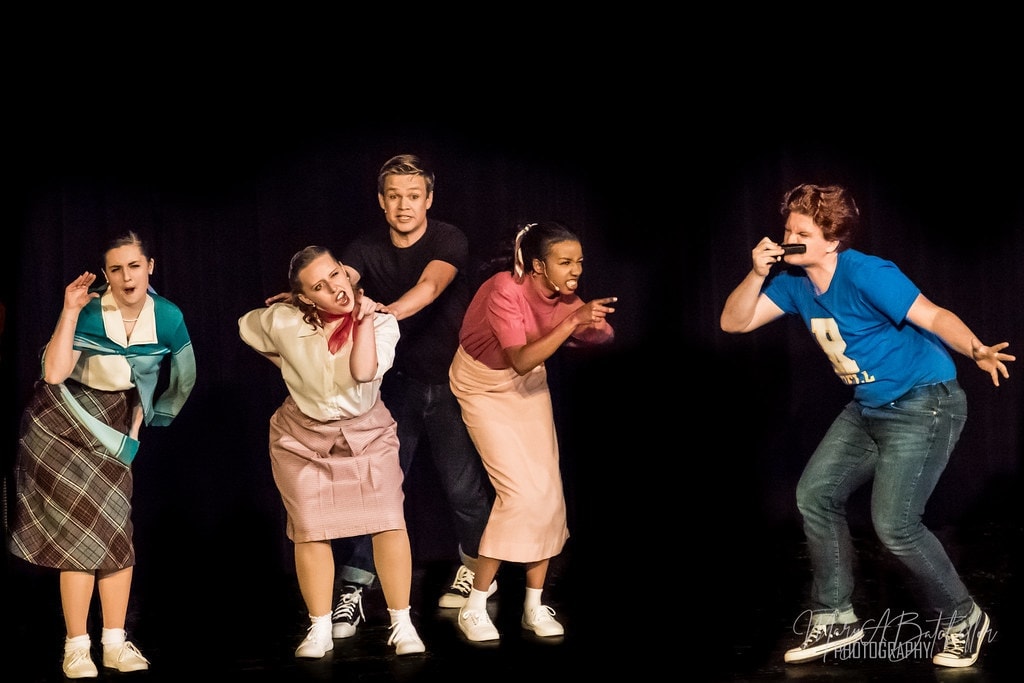 Grease — cast performing comedic number with microphone on dark stage