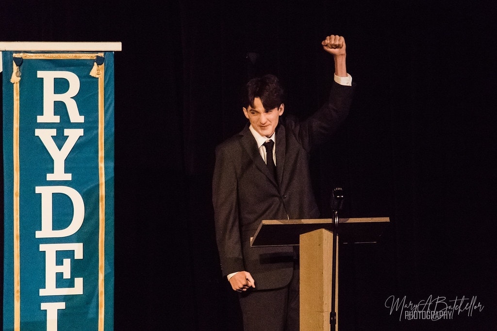 Grease — Danny at Rydell podium raising fist close-up portrait