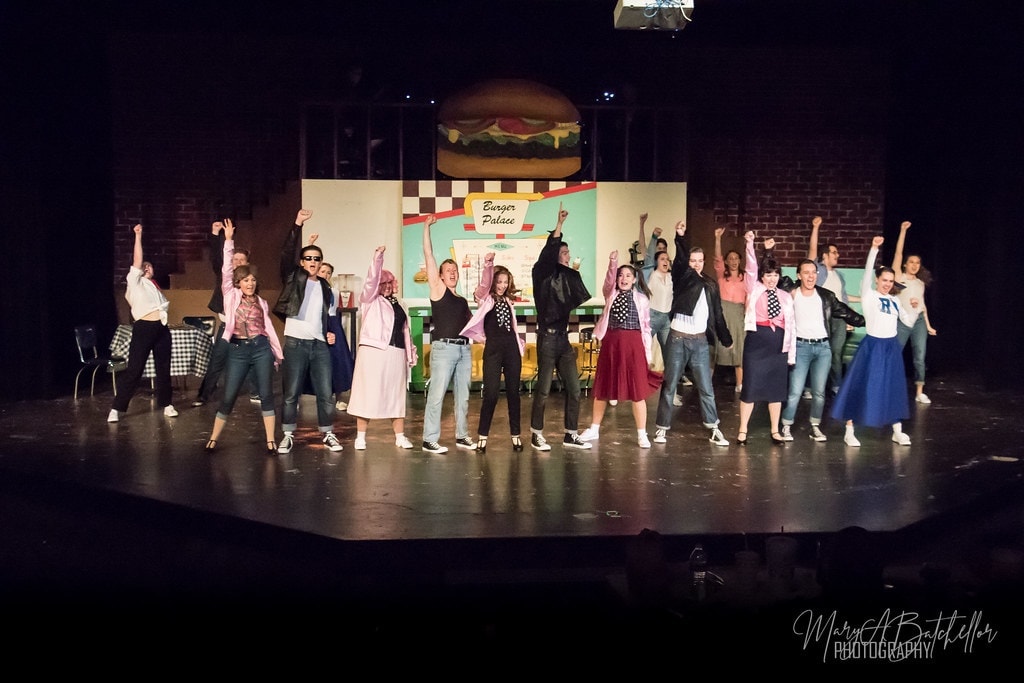 Grease — full cast with arms raised in front of Burger Palace set