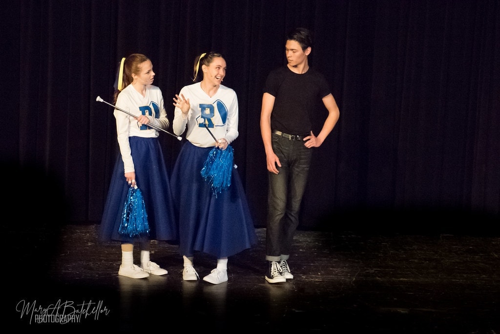 Grease — two Rydell cheerleaders with pom-poms and batons talking to Danny