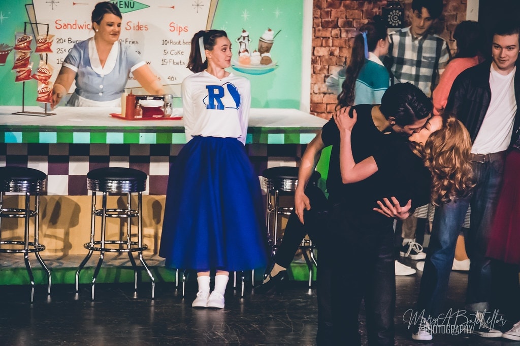 Grease — Sandy watches as Danny leans in to kiss a girl at the counter