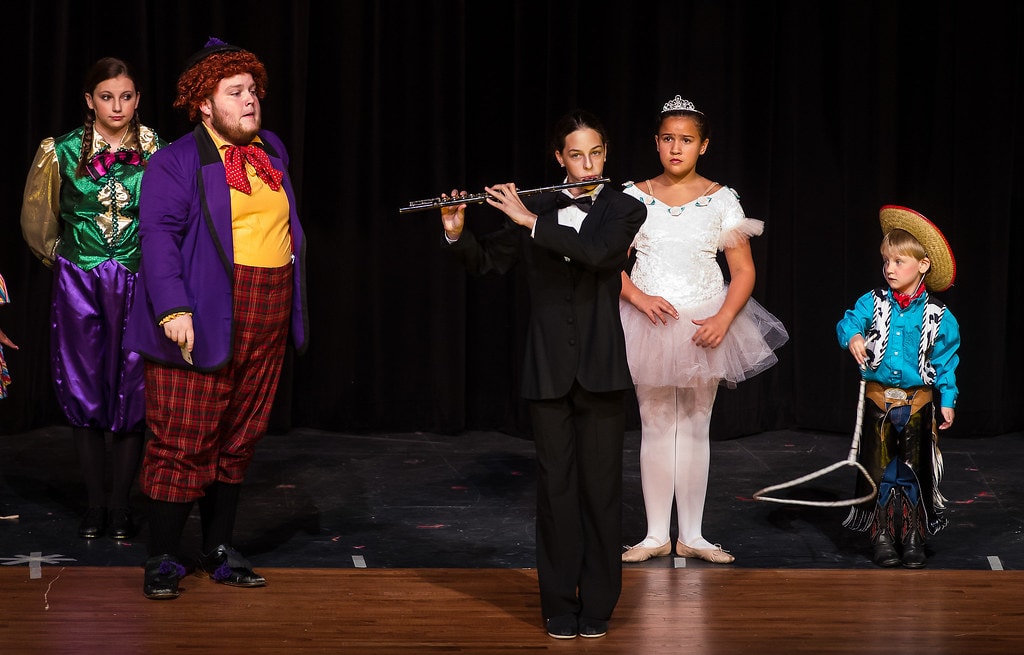 Gypsy — five children in eclectic vaudeville costumes stand on the bare black stage: a girl in a green-and-gold jester outfit, a boy in a purple plaid suit with red bow tie, a child in a black tuxedo playing flute, a girl in a white tutu with tiara, and a small boy in a teal cowboy shirt with sombrero and lasso