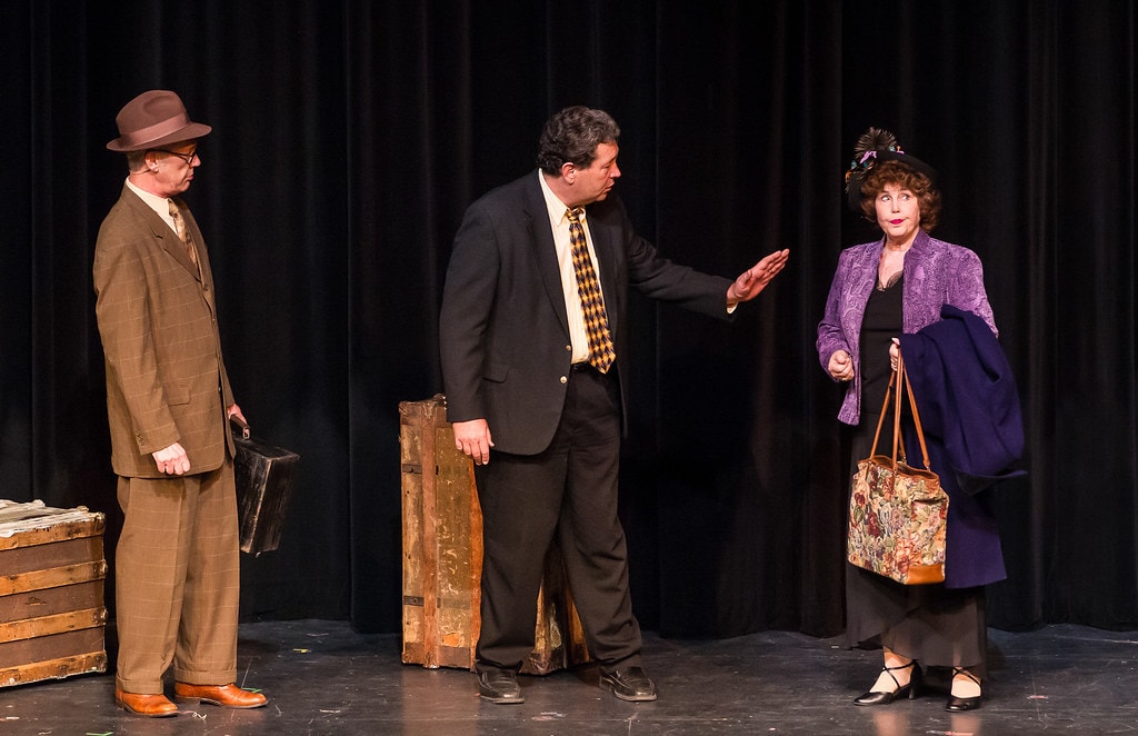 Gypsy — three adults in 1920s period suits stand before a black curtain backdrop: a man in a brown suit with briefcase at left, a man in a black suit with a gold-patterned tie gesturing with one hand toward a woman in a purple jacket and navy coat carrying a tapestry bag