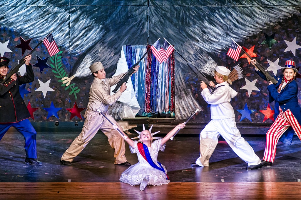 Gypsy — five performers in a patriotic vaudeville number: a marine, a soldier, a sailor, and a girl in Uncle Sam striped pants hold rifles aloft while a tiny girl in a white Statue of Liberty dress sits center stage with arms spread wide before a red, white, and blue star-spangled backdrop