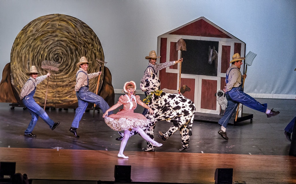 Gypsy — the farm act in full swing: the girl in the pink gingham dress dances with a performer in a dalmatian cow costume as four men in overalls and straw hats kick their legs high beside the giant hay bale and red barn set piece