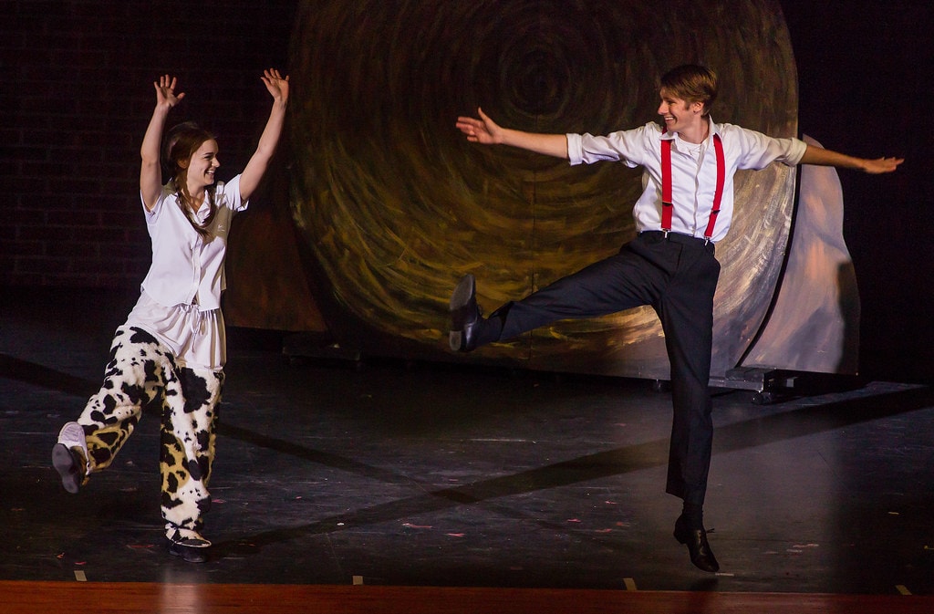 Gypsy — Louise in her cow-print pajamas kicks one leg high with arms spread wide as the young man in the white shirt and red suspenders mirrors her with a high kick and outstretched arms, both laughing joyfully before the giant painted hay bale backdrop
